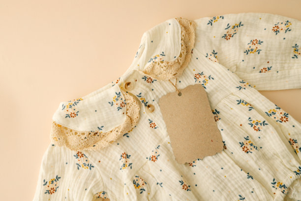 特写镜头展示了一件带有蕾丝细节的花卉婴儿裙，配有环保的牛皮纸标签，背景为柔和的米色。这张图片强调了可持续性和复古风格的婴儿服装设计。图片下载