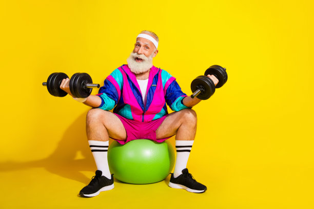 全身照片横幅，搞笑的老年运动员坐在健身球上举哑铃，穿着复古运动服，背景为黄色。图片下载