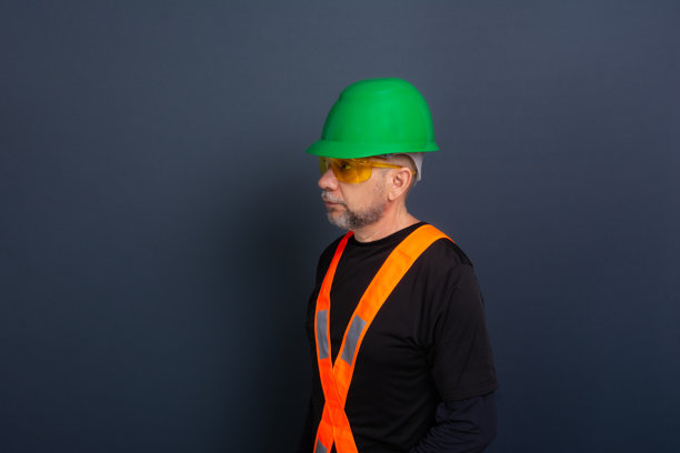 一名穿着绿色安全帽、反光背心和护目镜的工作人员。为照片摆姿势。背景为灰色。图片下载