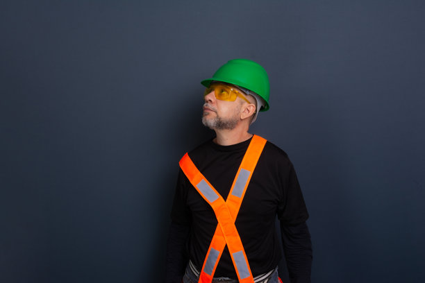 一名穿着绿色安全帽、反光背心和护目镜的工作人员。为照片摆姿势。背景为灰色。图片下载