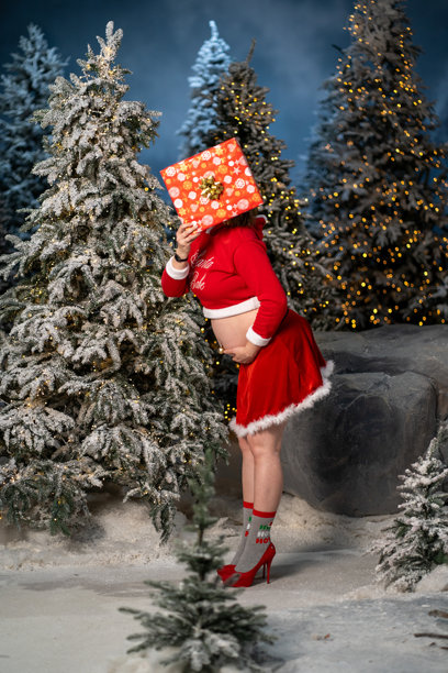 美丽的年轻孕妇穿着红色圣诞老人服装，站在冬季雪白的森林中，靠近圣诞树。圣诞快乐，新年快乐。节日怀孕。女人手里拿着礼物，准备送给未来的宝宝。图片下载