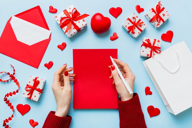 从上方拍摄的情人节装饰照片，女性手握着信件和信封，旁边有小礼物盒和心形装饰，背景为单色柔和色调，留有空白空间。图片下载