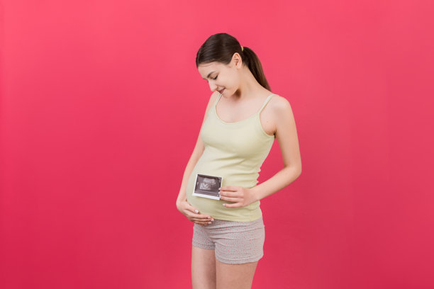 可爱的孕妇在彩色背景前摆姿势，手持宝宝超声波照片。孕期、妇科、医学检查、母婴健康的概念。图片下载