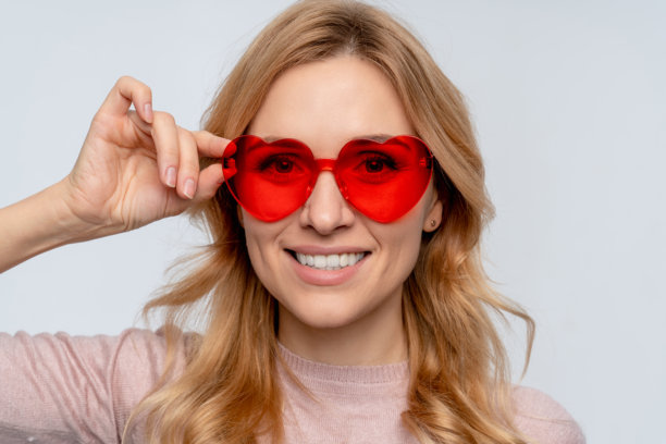 特写照片：快乐可爱的年轻女性戴着时尚的红色心形太阳镜，微笑着看向镜头，站立在白色背景前。图片下载