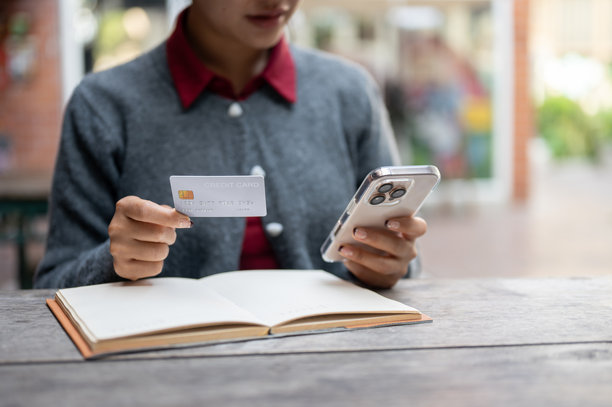一张裁剪过的图片，展示了一位女性坐在户外桌子旁，手中拿着智能手机和信用卡。图片下载