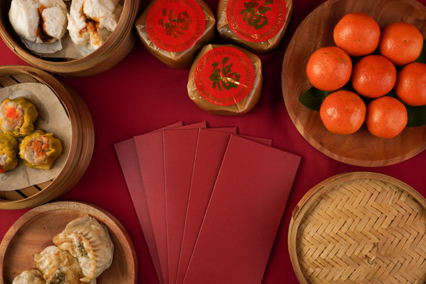 一张平铺的照片，展示了一张红色桌布，上面摆放着各种传统的中国新年菜肴，如点心、橙子、年糕和红包。（汉字“福”代表着财富）图片下载