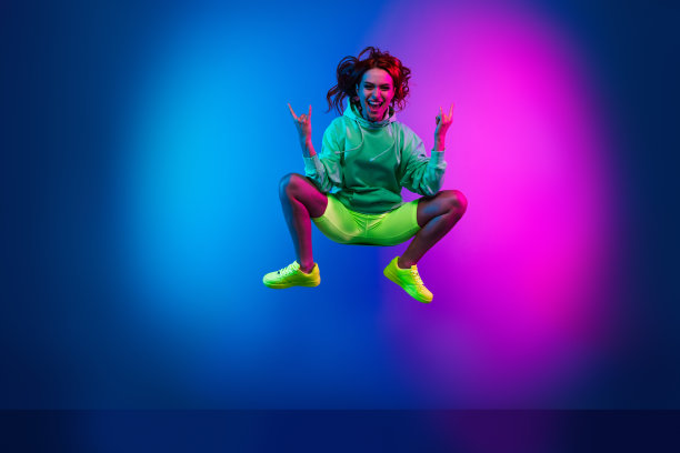 穿着卫衣的快乐酷女孩在空旷的霓虹粉色背景下高高跳起，做出摇滚手势的全身照片图片下载