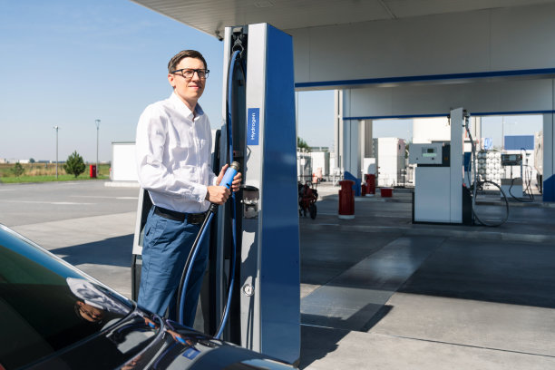 男子穿着白衬衫和蓝色牛仔裤，在路边的氢气加注站握着氢气加注枪。为汽车加注氢燃料。阳光明媚的日子。图片下载