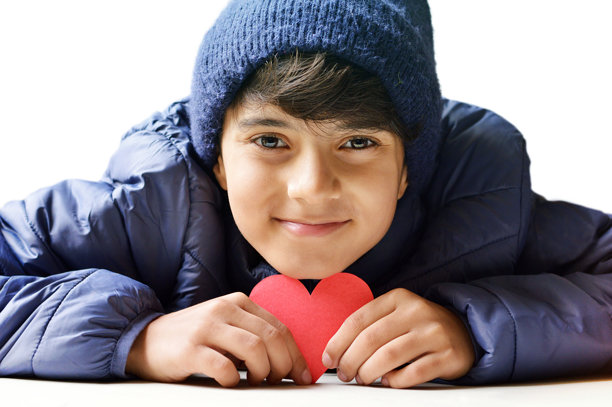 一个快乐微笑、嘴唇紧闭的12岁可爱男孩，穿着蓝色夹克和毛线帽，手持纸剪心形图案，靠近脸颊和下巴；情人节相关图片，孤立在水平白色背景上。图片下载