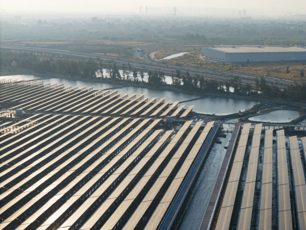 海边太阳能电池板发电厂的航拍照片，远处是工业厂房。图片下载