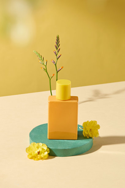 这张香水广告照片展示了一个橙色瓶子的模型，放置在一个绿色圆形底座上。瓶子周围摆放着两朵黄色花和一小枝。背景是浅黄色的。图片下载