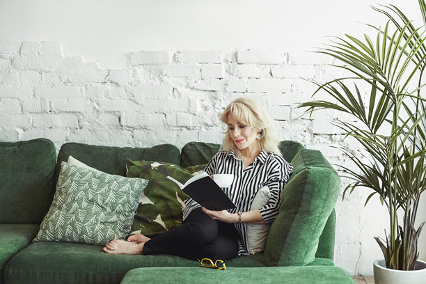吸引人的时尚六十岁女性的自然照片，悠闲地专注于爱好，享受经典文学，舒适地坐在大沙发上，手里拿着书和一杯热饮。图片下载