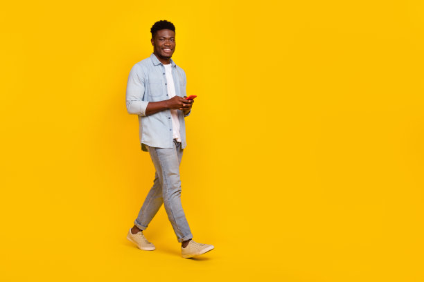 全身照片，千禧一代男士走路时使用智能设备与博客粉丝沟通，背景为明亮的颜色。图片下载