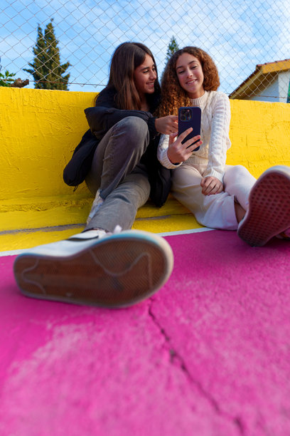 两个少女坐在五彩斑斓的矮墙上，正在用智能手机查看照片。图片下载