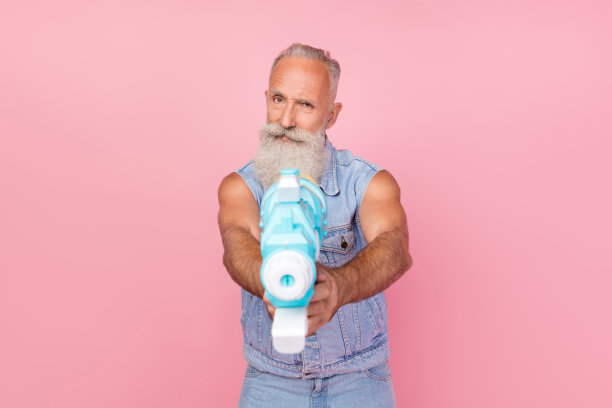 穿着牛仔裤和马甲的时髦酷炫退休男士，手持水枪对着你，背景为粉色。图片下载