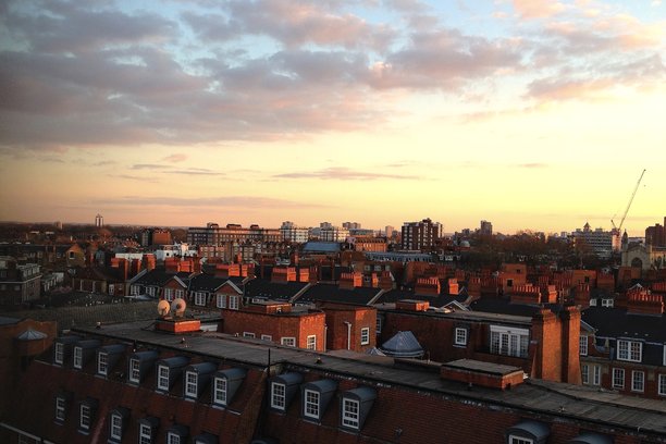 一幅城市风景照片，捕捉了日落时分伦敦的屋顶。画面中有红砖烟囱、经典建筑，以及散布的云朵映衬下的橙色天空。图片下载