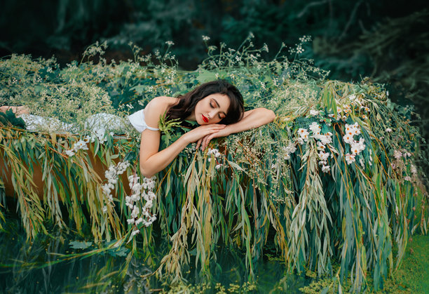 艺术照片：美丽的睡眠女性在船上休息，周围有柳树枝叶和白色花朵。希腊女神水妖精灵女孩闭着眼睛享受自然河流的宁静。幻想童话公主。图片下载