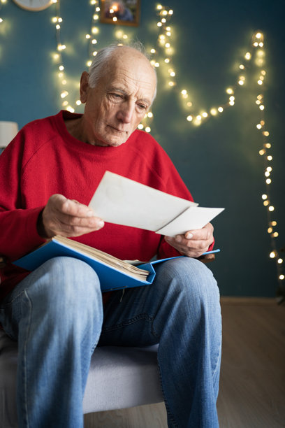 一位老年男子在圣诞前夕独自坐在扶手椅上，翻看着旧相册，回忆年轻时的照片。图片下载