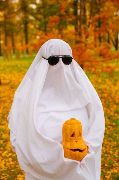 穿着白色床单的女人手持南瓜灯，背景是秋季森林。万圣节服装。竖版照片。图片下载
