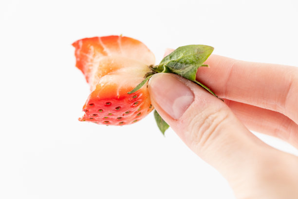 在白色照片背景上，纤细的女性手握着一半被咬过的成熟草莓，草莓的尾部，健康的维生素，时令，夏季水果，维生素C图片下载