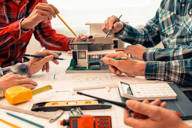一群多民族的男性和女性建筑师及工人围绕办公桌站立，查看桌上的纸质蓝图和房屋设计图。特写照片。图片下载