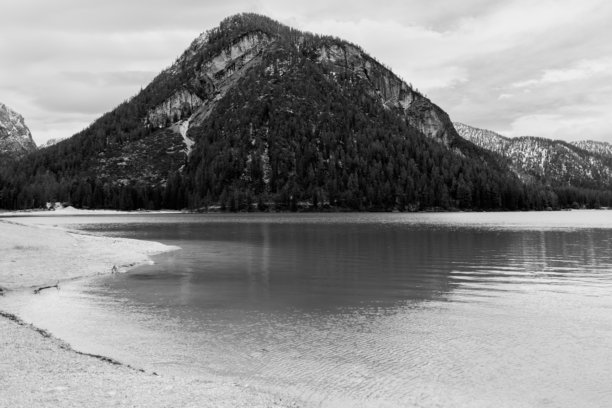 意大利多洛米蒂的布拉耶斯湖，湖水平静，沙滩岸边密集的常青森林和陡峭的山峰在阴云密布的天空下（黑白照片）图片下载