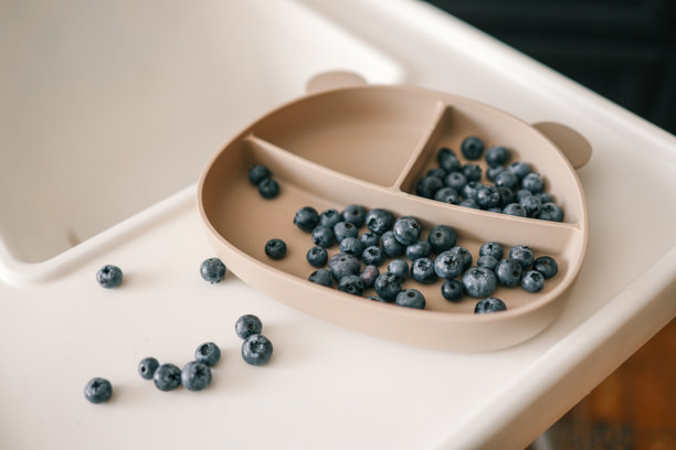 高脚椅托盘上的硅胶吸盘盘中放着蓝莓，周围散落着一些。现代厨房环境，简约的幼儿用餐，婴儿主导的辅食添加和健康零食概念。图片下载