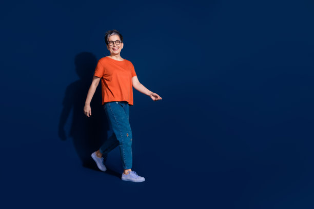 穿着时尚橙色服装的可爱退休女士在深蓝色背景下走路的全身照片图片下载