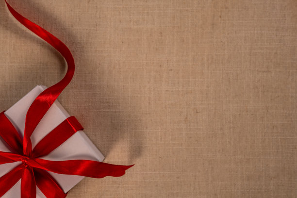 米色纹理背景，左上角有一个用红色丝带蝴蝶结系好的白色礼品盒，审美风格的模拟照片背景图片下载