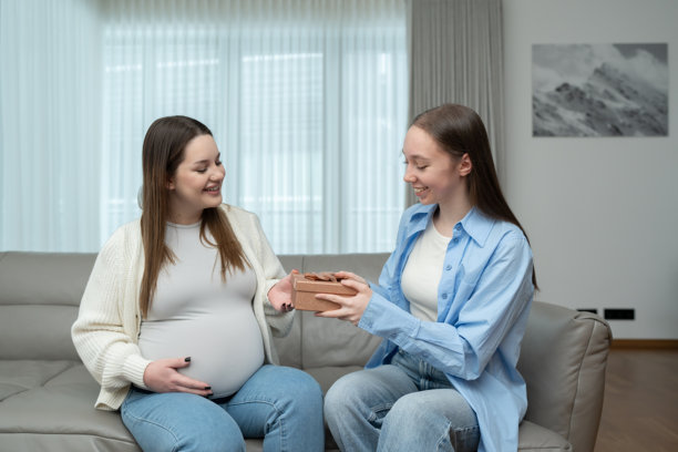 微笑的孕妇坐在舒适的家中沙发上，接受女性朋友送来的婴儿礼物。母性、庆祝、友谊、期待和情感联系的概念。图片下载