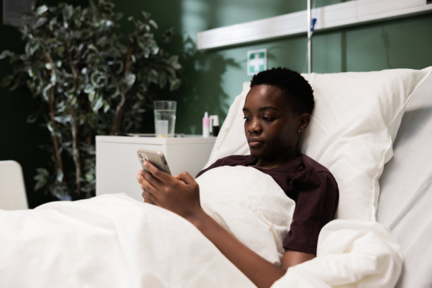 年轻女性躺在医院病床上，手中拿着手机，可能在翻阅熟悉的消息或照片中寻找安慰。图片下载