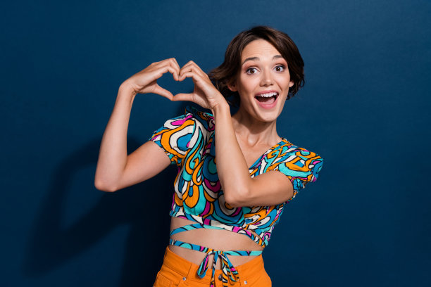 漂亮年轻女孩的照片肖像，展示心形手势，穿着时尚的喇叭裤牛仔服，背景为深蓝色。图片下载