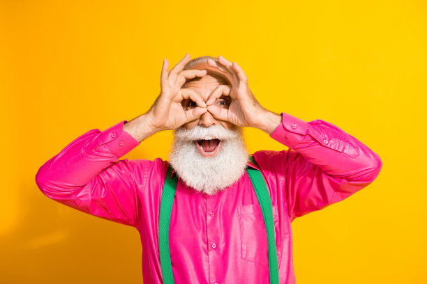 时尚老绅士的照片肖像，做出使用双筒望远镜的手势，穿着华丽的吊带裤，背景为明亮的黄色。图片下载