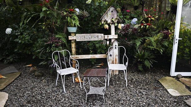 户外花园休息区，配有白色金属椅子、木质桌子、标有“花卉”的乡村风格木牌，四周环绕着郁郁葱葱的绿植和鲜艳的花朵，地面为碎石铺成。图片下载