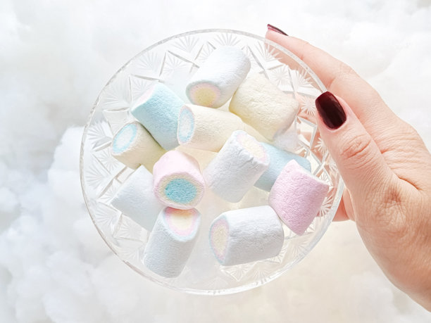 一只女性的手握着一个装有棉花糖的玻璃糖果碗，背景是柔软的雪白棉花糖云朵。棉花糖的宣传光影照片。图片下载