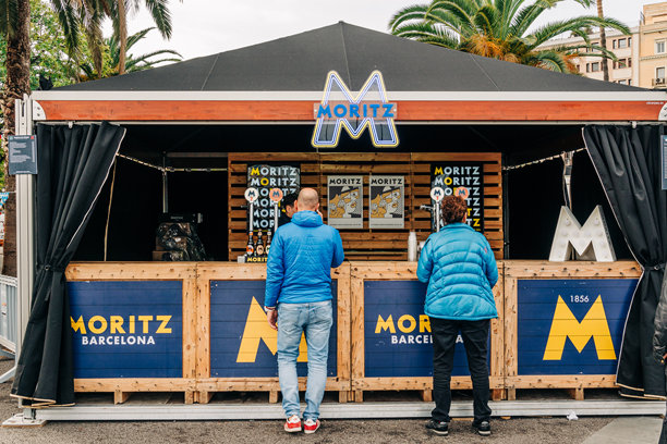 一名穿着蓝色夹克的男子站在巴塞罗那的Moritz啤酒摊位前，摊位上有品牌标识、酒龙头和一个木质柜台，搭在帐篷下。图片下载