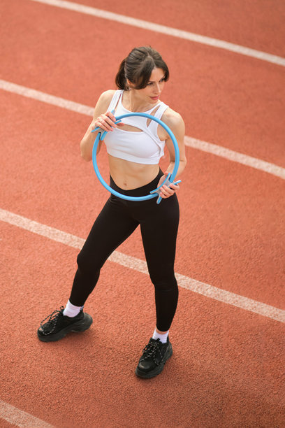 一位健康的女性在跑道上将普拉提环举过头顶，坚定的表情彰显了她的专注。这张图片展示了使用健身工具来增强锻炼效果。图片下载