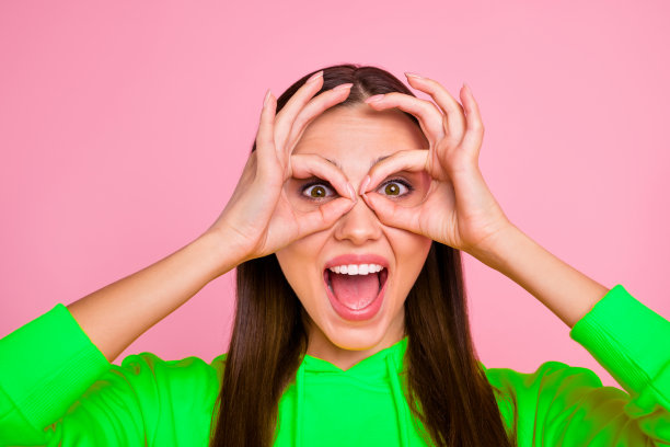 特写照片，时尚、快乐、可爱、迷人、吸引人的女友穿着鲜艳的黄色毛衣，双手做出“OK”手势环绕在眼睛周围，背景为粉色柔和色调。图片下载