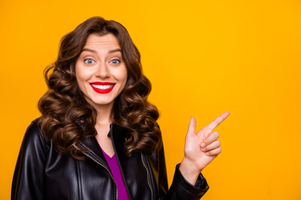 特写照片，漂亮的卷发女士用手指指向空白处，穿着皮夹克，背景为黄色，表示低价。图片下载