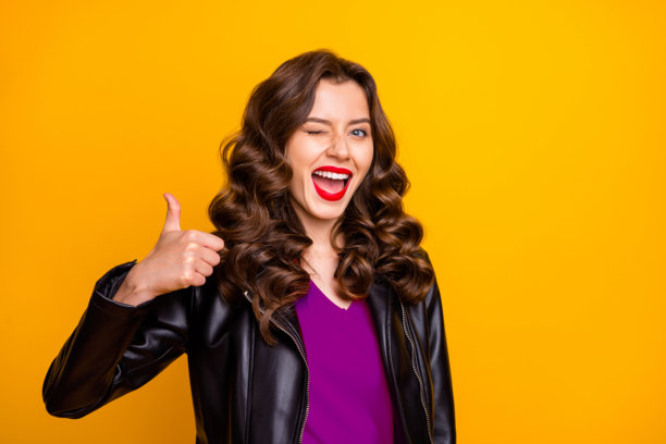 特写照片：一位女士嘴唇涂红，竖起大拇指，眨眼，穿着皮夹克，背景为黄色。图片下载