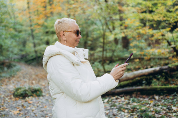 50岁女性游客在户外使用智能手机。老年女性在自然中徒步旅行。美丽的金发女性在秋季风景中漫步。旅行、科技与人群的横幅。高质量照片图片下载