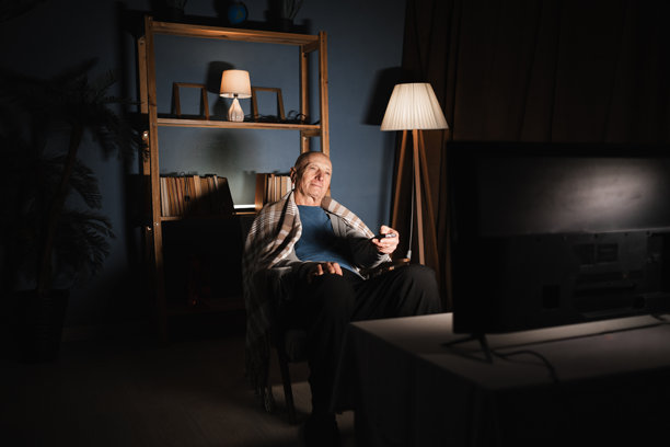 孤独的退休老人晚上独自在家坐在椅子上看电视，脸上带着微笑，手中握着遥控器切换频道。图片下载