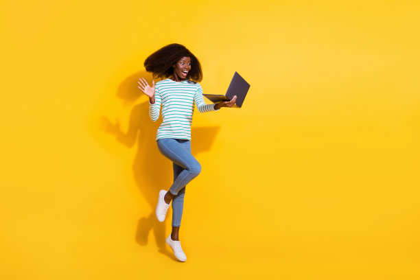 兴奋惊讶的女孩在黄色背景上跳跃，凝视着无线笔记本电脑的全身照片图片下载