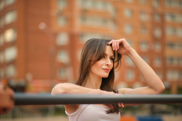 一位健康的女性微笑着倚靠在健身器材上，放松的姿态散发出自信。这张图片将身体健康与心理健康以及城市生活联系在一起。图片下载