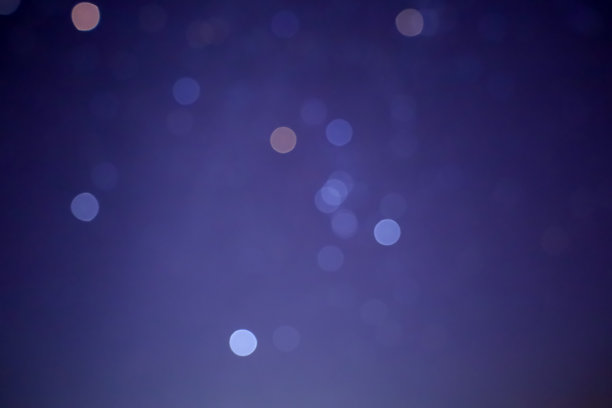 照片展示了在黑暗背景上模糊的光点。这些光点呈圆形，具有不同的白色和橙色阴影。它们在图像中杂乱无章地排列，形成了散景效果。背景呈深蓝紫色。图片下载