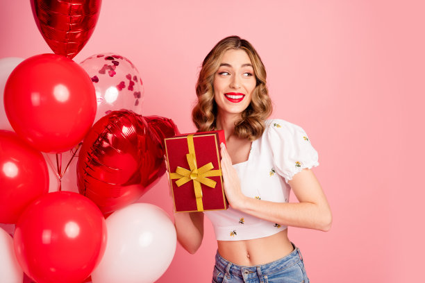 可爱的年轻女性的照片肖像，手持气球和礼物，目光投向空旷的空间，穿着时尚的白色服装，发型精致，背景为粉色。图片下载
