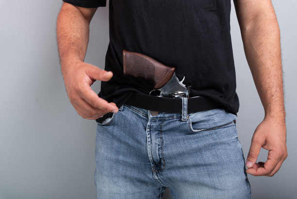 穿着黑色衬衫和牛仔裤的射击教练，腰间挂着左轮手枪，背景为灰色。体育练习。图片下载