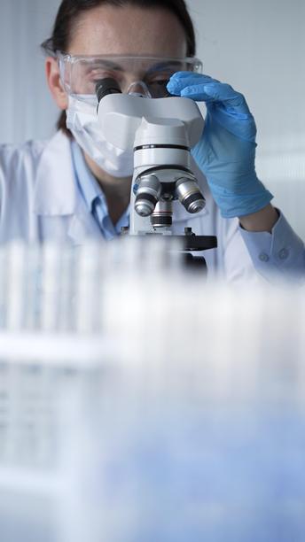 穿着蓝色手套、护目镜和医用口罩的女科学家正在现代实验室中使用显微镜。低调照明的照片。科学与医学图片下载