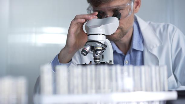 穿着实验室外套和防护眼镜的男性科学家在现代实验室中使用显微镜进行研究，肖像视图。低调照明照片。科学与医学概念图片下载