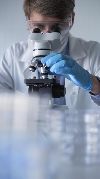 穿着蓝色手套、护目镜和医用口罩的男性科学家研究员正在现代实验室中使用显微镜，肖像视图。低调照明照片。科学与医学概念。图片下载